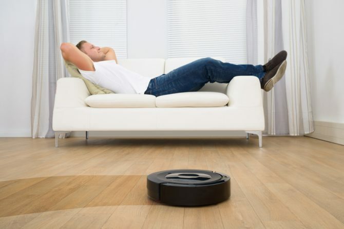 Žmogus atsipalaiduoja ant sofos su robotu dulkių siurbliu ant kietmedžio grindų